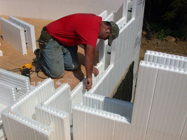 A worker in a red shirt installs and levels white CelBlox ICF blocks at a wall corner, showing the interlocking ridges near a window opening.