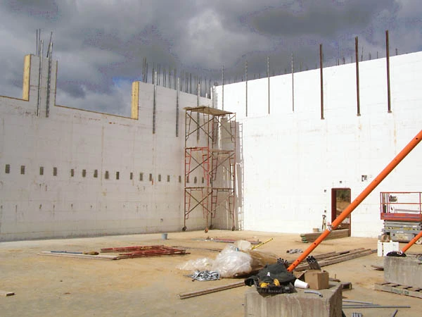 Interior view of tall white CelBlox insulated concrete form walls for the Flaherty School gymnasium, with rebar dowels awaiting the next pour, a scaffold access tower, and storm clouds overhead.