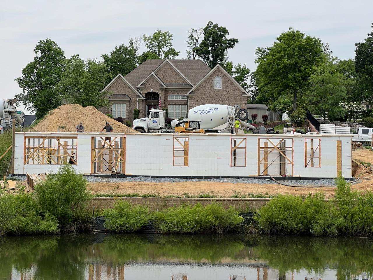 A residential ICF home build with white insulated concrete form walls and framed window openings. A concrete mixer truck is parked beside the site with a brick house and pond in the background.