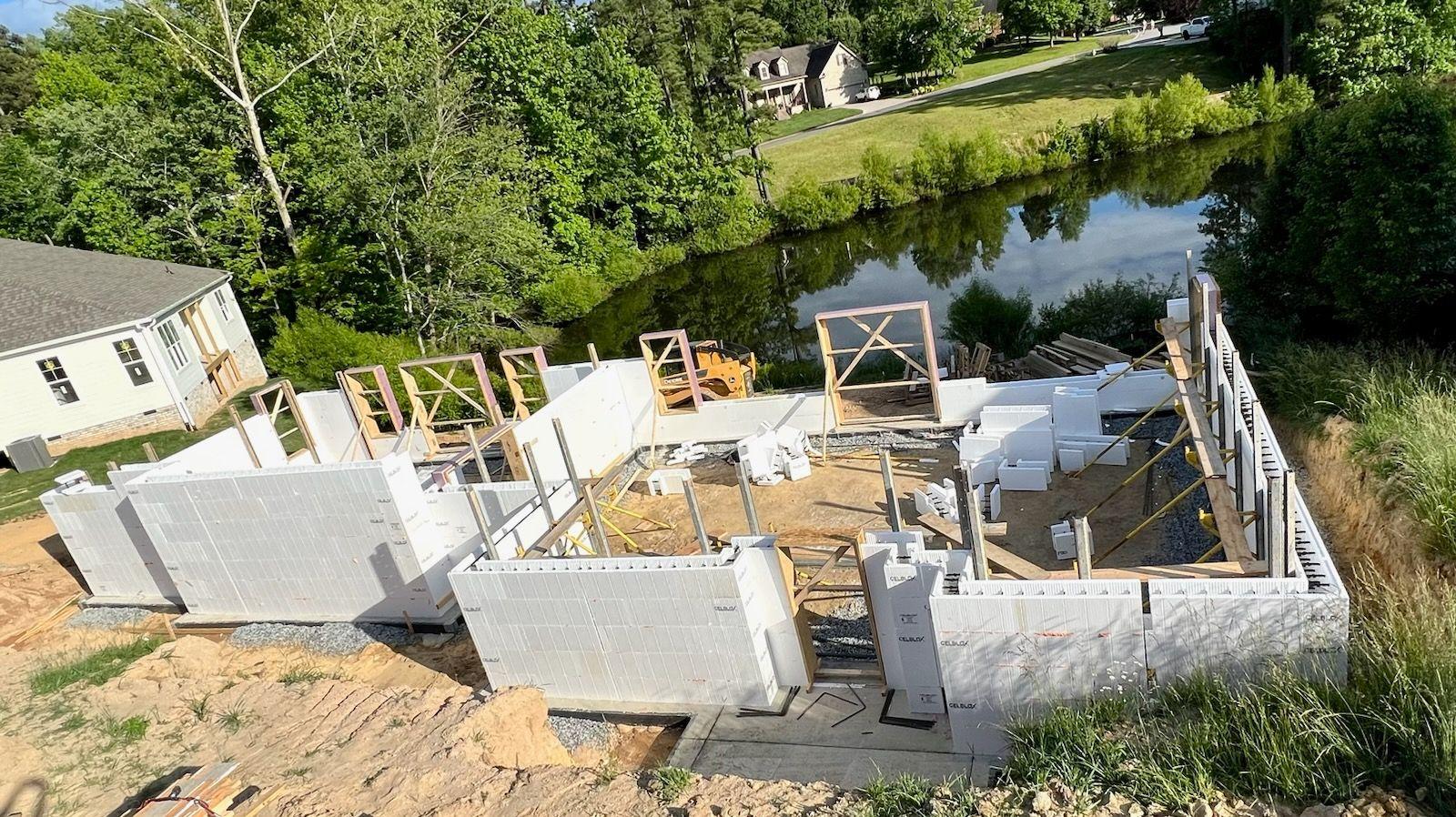 ICF wall panels being assembled for a residential ICF home build along a riverbank, with wood bracing supporting the insulated concrete form walls and neighboring houses visible across the water.