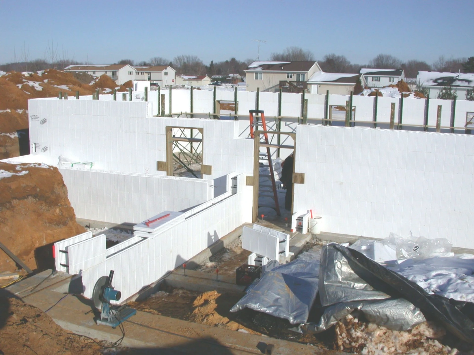 ICF construction site in winter, with insulated concrete form foundation walls being built near a neighborhood of homes with snow-covered rooftops in the background.