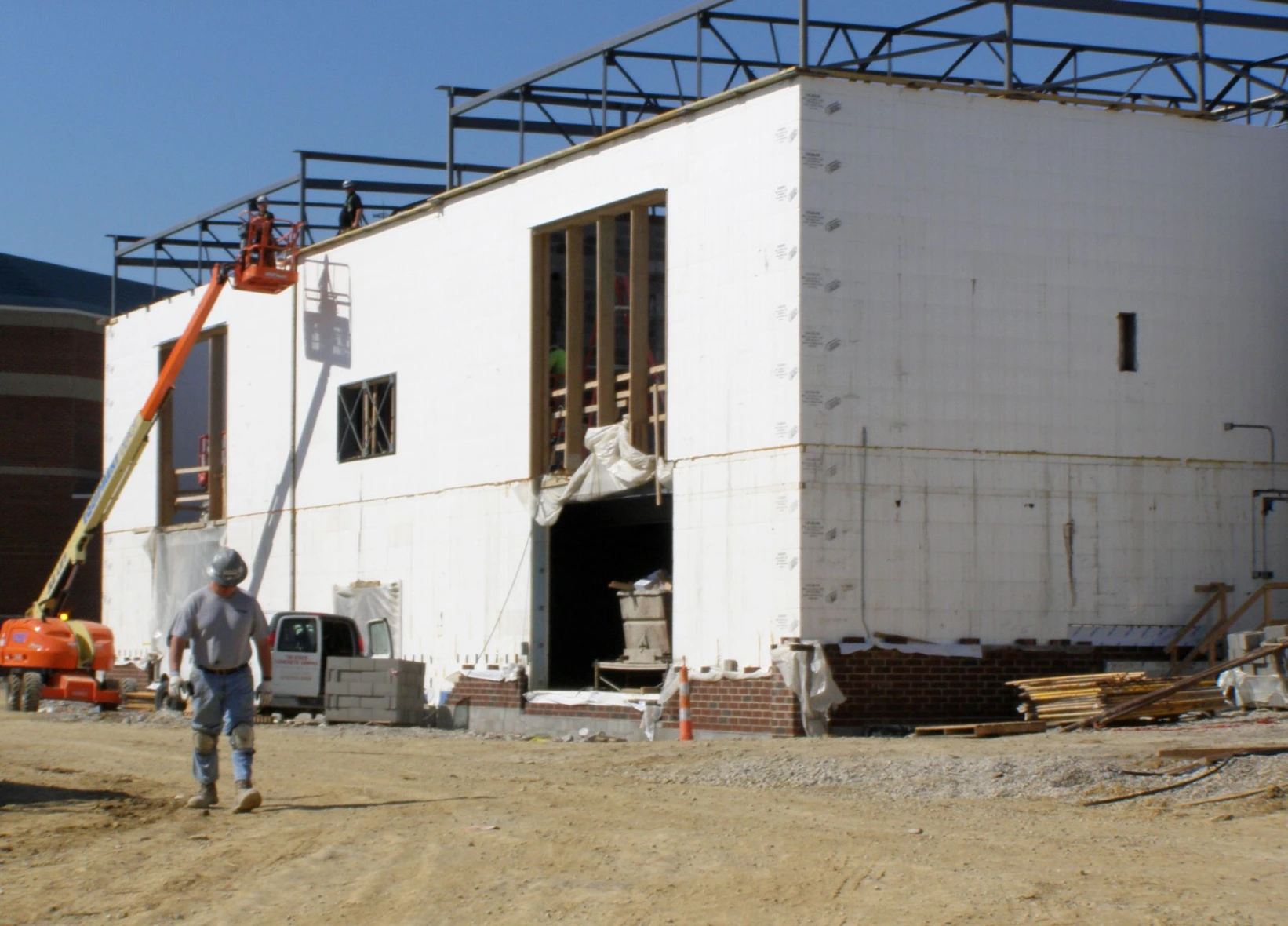 Corner of a commercial ICF building near completion, with a worker in a hard hat walking past the white insulated concrete form walls, an orange scissor lift, and brick foundation detailing.