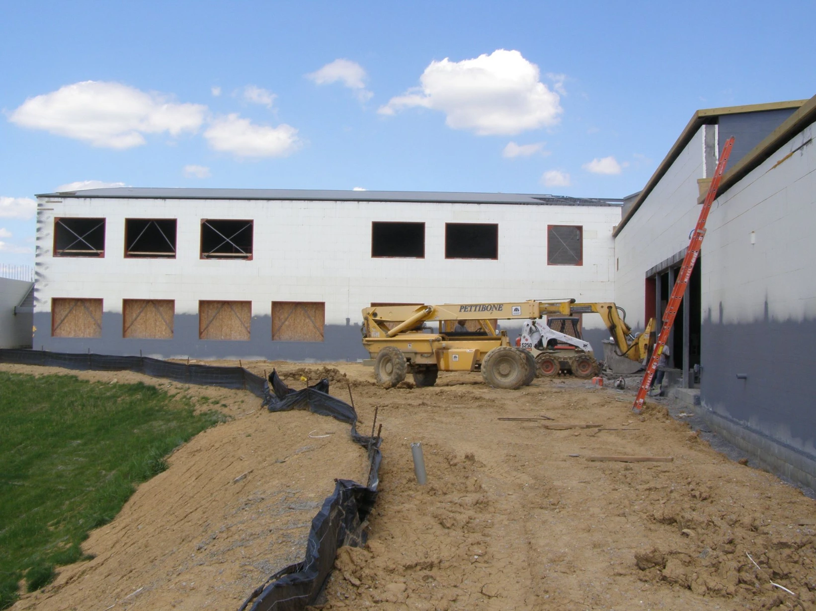 Long commercial ICF building with white insulated concrete form walls, multiple framed window openings, a yellow skid steer loader, and landscape fabric along the foundation trench.