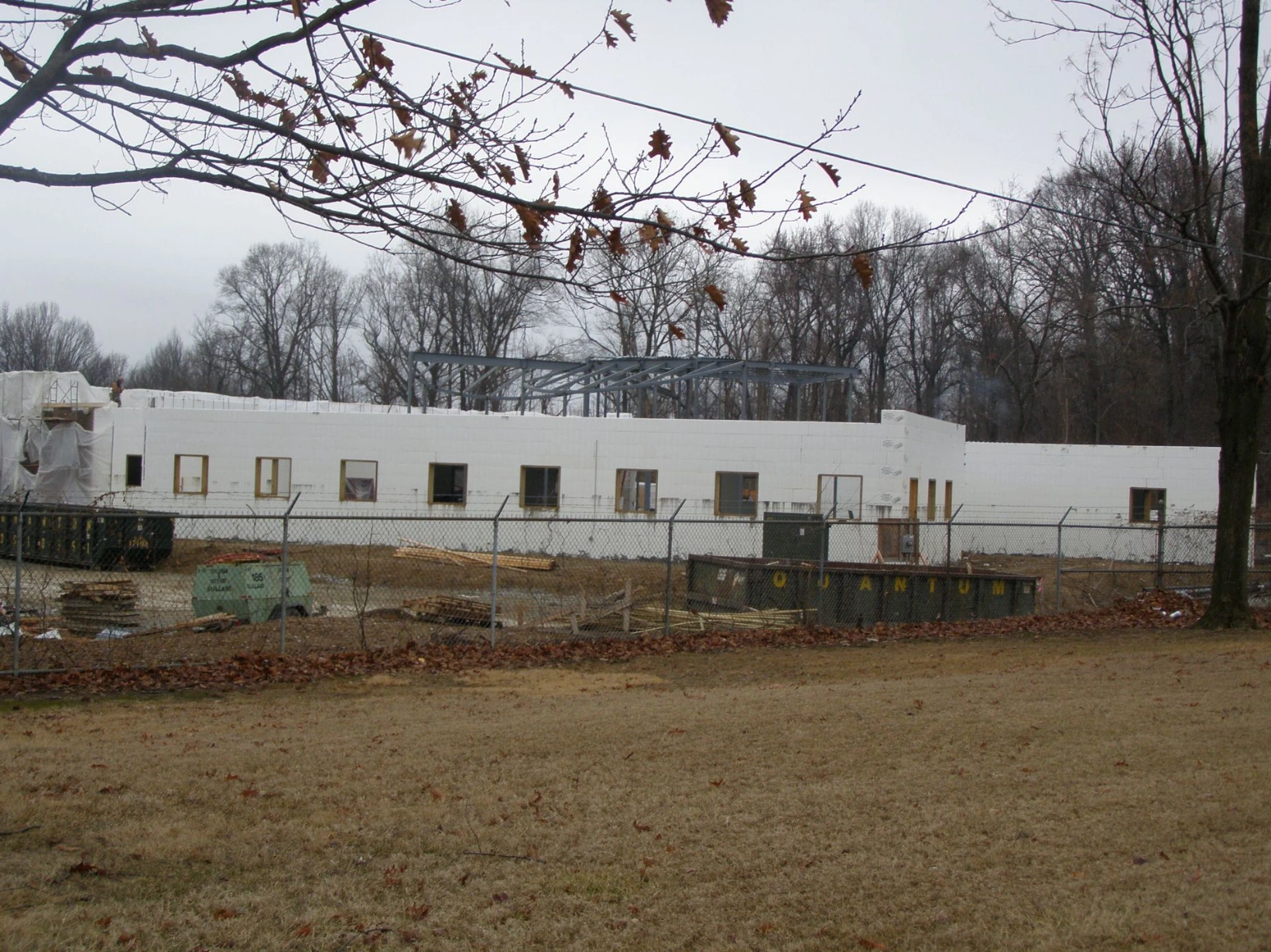 Long commercial ICF building with white insulated concrete form walls, a fenced construction site, dumpsters, and steel roof trusses being erected on one end.