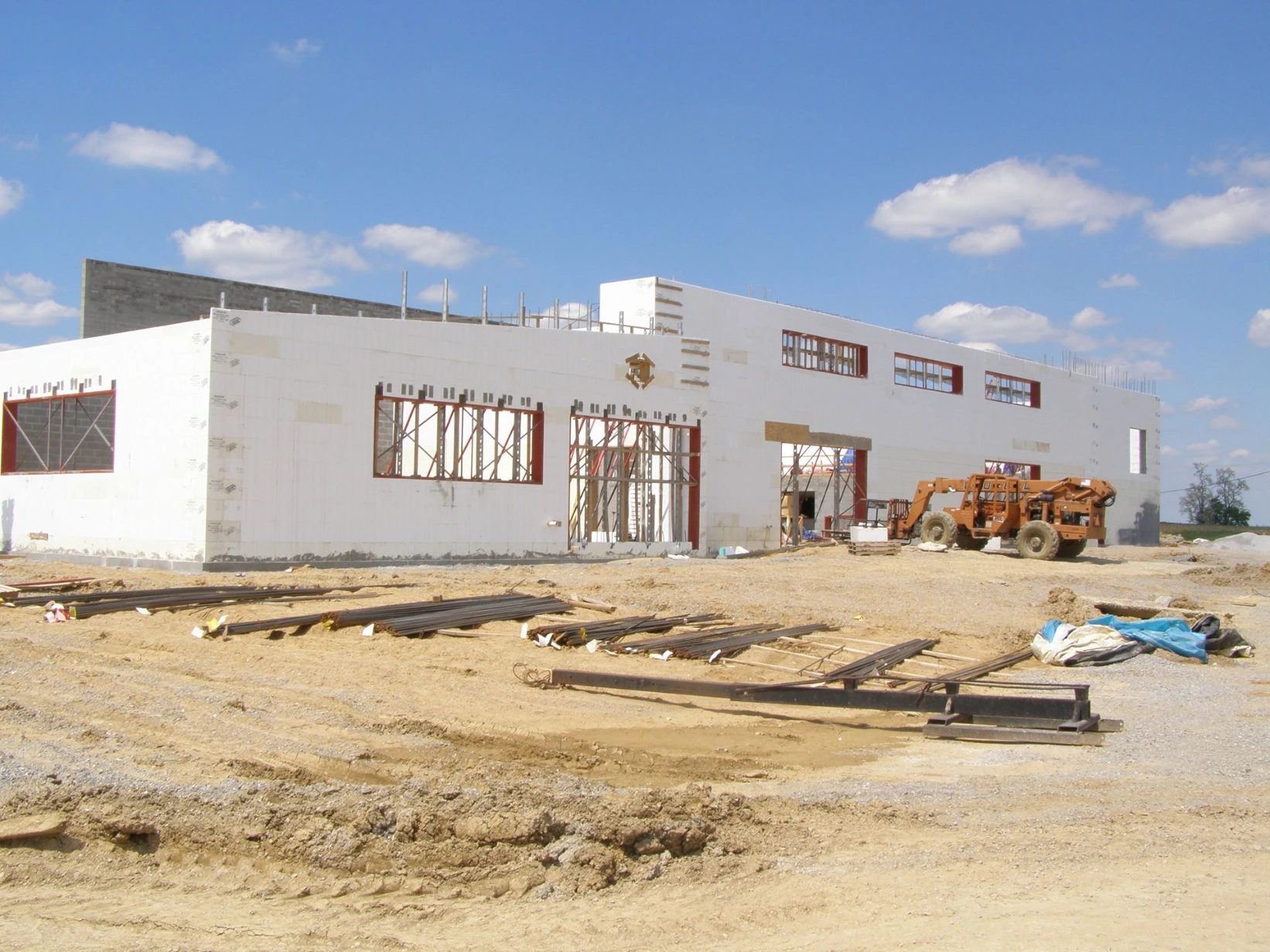 Long commercial ICF building with a wide bay door opening, red-framed windows, an orange telehandler, and steel rails staged on the ground at a rural job site.